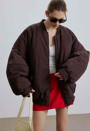 Kobieta w oversize'owej, ciemnobrązowej kurtce puchowej, białym prążkowanym topie na ramiączkach, czerwonej spódnicy, czarnych okularach przeciwsłonecznych, trzymająca beżową torebkę, patrząca w dół.