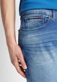 Blue denim jeans with a faded texture, featuring a front pocket and detailed stitching, including a red accent on the pocket lining.