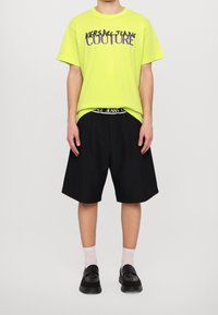 Neon žluté bavlněné tričko s grafikou "VERSACE JEANS COUTURE", spárováno s černými šortkami ke kolenům a černými botami. Jednoduché pozadí.
