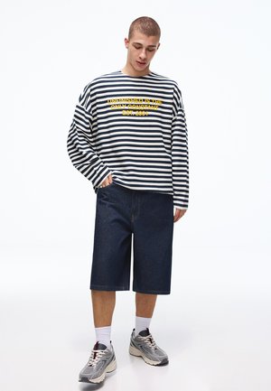 Mladi moški, oblečen v črno-belo črtasto majico z dolgimi rokavi in rumenim napisom, temne kratke hlače do kolen, bele nogavice in sive superge.