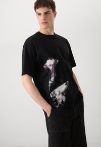 Črn majica iz bombaža, ki ima grafično zasnovo z meduzami v beli in vijolični barvi. Ohlapen kroj z okrogllim izrezom.