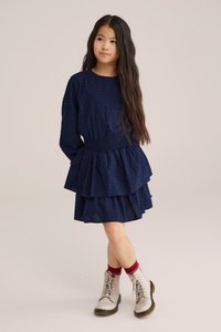 Sötétkék ruha strukturált mintával, hosszú ujjakkal és réteges, fodros szoknyadizájnnal. Világos színű fűzős csizmával és piros zoknival párosítva.
