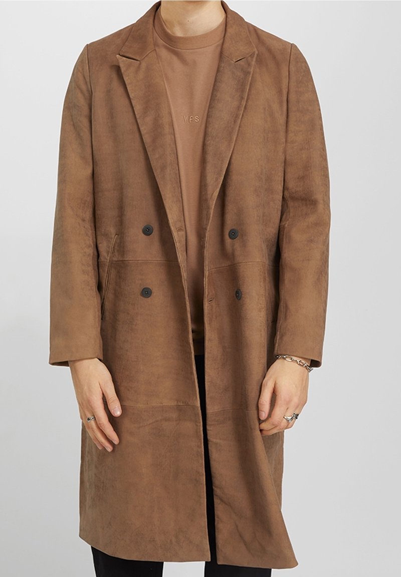 Osoba nosící hnědý semišový dlouhý kabát s černými knoflíky přes sladěnou hnědou košili, s prsteny a náramkem na rukou.