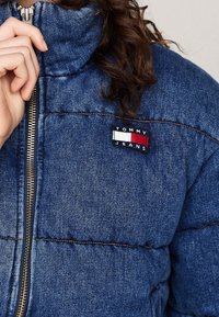 Modrá džínová péřová bunda s vysokým límcem, opatřená zipovým uzávěrem a obdélníkovou nášivkou s logem "TOMMY JEANS" v červené, bílé a černé barvě.