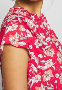 Bloemige blouse in roze met witte en koraalkleurige bloemen. Kenmerkt zich door een gerimpelde halslijn, korte mouwen en een lichte, gestructureerde stof.