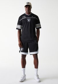 Maillot en maille noire avec des rayures blanches et argentées, arborant le logo des Raiders. Associé à un short noir et des baskets blanches. Casquette de couleur claire.