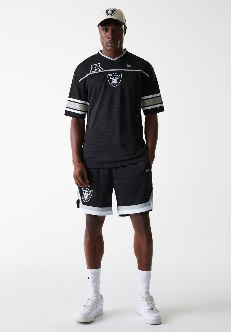 Maillot en maille noire avec des rayures blanches et argentées, arborant le logo des Raiders. Associé à un short noir et des baskets blanches. Casquette de couleur claire.