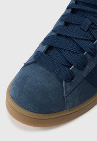 Κοντινό πλάνο μπλε σουέτ αθλητικού παπουτσιού με χονδρές ναυτικές μπλε κορδόνια και ελαφρώς καφέ λαστιχένια σόλα σε λευκό φόντο.