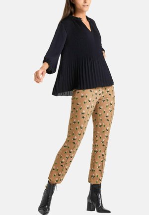 Vrouw met een losse zwarte geplooide blouse, beige broek met groene en zwarte bijenpatronen, en zwarte enkellaarsjes met hak, staand tegen een witte achtergrond.