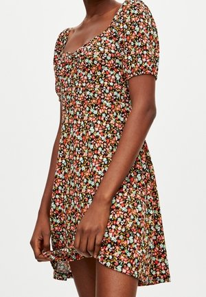 Femme portant une robe courte à imprimé floral avec des manches bouffantes et un décolleté carré, tenant l'ourlet de la robe.