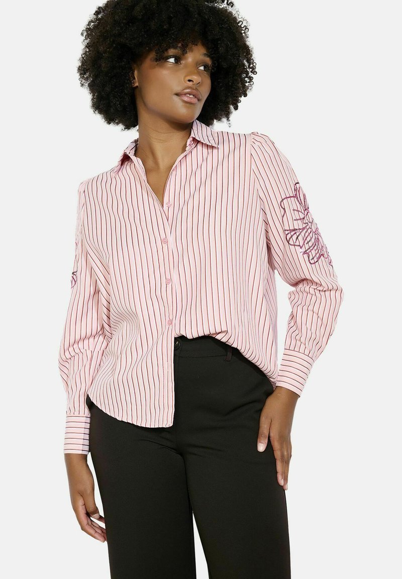 Ružová košeľa s vertikálnymi pruhmi, s dlhými rukávmi, so sýtočerveným kvetinovým vyšívaním na ľavom ramene, nosená s čiernymi nohavicami.
