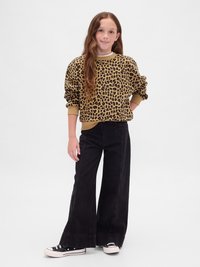 Ragazzina con capelli lunghi che indossa un maglione in stampa leopardata, pantaloni neri a gamba larga e scarpe sportive bianco e nere, che sta in piedi su uno sfondo semplice.