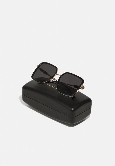 Juodų akinių nuo saulės rėmas su tamsiais objektyvais ir auksinėmis detalėmis, padėti ant blizgaus juodo kietojo dėklo su iškilniu "VERSACE" logotipu.