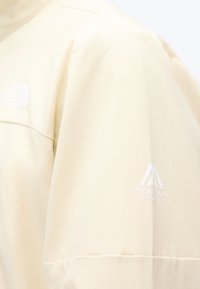 Lysebeige jakke med Mountain Athletics-logo på ærmet og et subtilt hvidt logo på brystområdet.