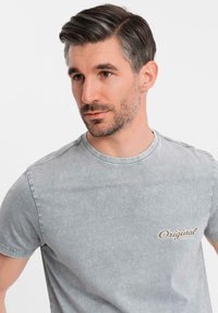 Camiseta de algodón gris con cuello redondo, mangas cortas y un pequeño gráfico de "Original" en color beige en el lado izquierdo del pecho. Textura suave.