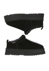 Paar schwarze Slip-on-Winterschuhe mit dicken, profilierten Sohlen und weichem Innenfutter, seitlich dargestellt, wobei ein Schuh umgedreht ist, um die Sohle zu zeigen.
