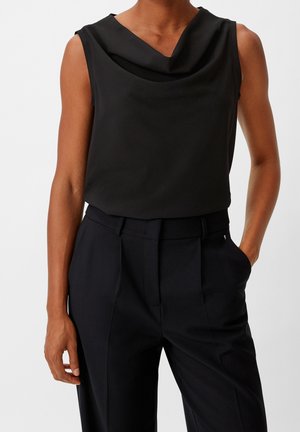 Femme portant un haut noir sans manches avec un col drapé et un pantalon noir taille haute, une main dans la poche.