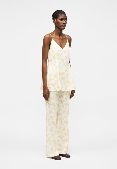 Γυναίκα στέκεται φορώντας λευκή αμάνικη τουνίκ με V-λαιμόκοψη και ασορτί παντελόνια με φαρδιά πόδια με ανοιχτό κίτρινο λουλουδάτο μοτίβο και σκούρα παπούτσια.