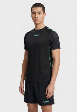 Jeune homme portant des vêtements de sport noirs et foncés à motifs avec le logo vert "hummel", debout devant un fond clair uni.