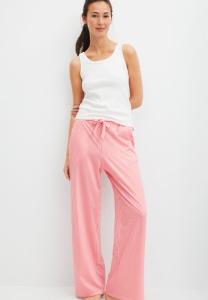 Vrouw die tegen een witte muur staat, gekleed in een wit mouwloos shirt en losse roze broek met verticale witte strepen en een taille met trekkoord.