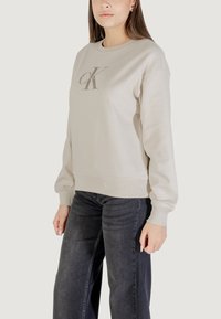 Sudadera beige hecha de suave algodón con cuello redondo, puños y dobladillo de canalé, que presenta un logo de Calvin Klein en plata oscura en la parte delantera.