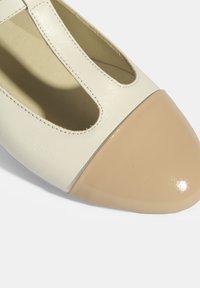 Lederschuh mit einem cremefarbenen Obermaterial, Ausschnitt-Details und einer glänzenden beigen Zehenkappe. Mit gestickten Akzenten entlang der Kante.