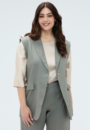 Uśmiechnięta kobieta z długimi brązowymi włosami, ubrana w jasnobeżową bluzkę oraz pasujący do niej jasnoszaro-zielony bezrękawnik i spodnie na białym tle.