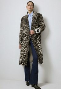 Femme portant un long manteau imprimé léopard avec un patch de cygne noir sur la manche, une chemise légère à boutons bleu clair, un jean évasé noir et des bottes noires.