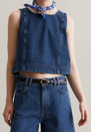 Osoba nosząca bezrękawnik typu cropped z denimu, dżinsy z wysokim stanem z czarnym paskiem oraz niebieską wzorzystą apaszkę na szyi na jednolitym tle.