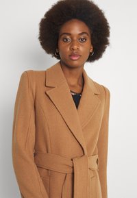Cappotto di lana color cammello con vita cintrata, rever a scollo e finitura testurizzata. Presenta spalle strutturate e tasche laterali.