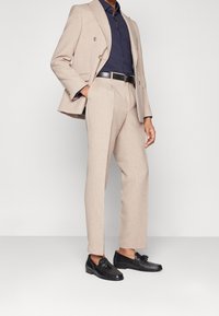 Chaqueta de traje de doble botonadura en beige claro y pantalones a juego, combinado con una camisa azul oscura y mocasines negros con detalle de borlas.