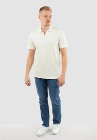 Weißes strukturiertes Poloshirt mit einem Reißverschlusskragen, kombiniert mit blauen Jeans und weißen Sneakern. Das Model steht vor einem neutralen Hintergrund.