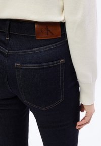 Σκούρο μπλε τζιν παντελόνι με κίτρινη ραφή, που διαθέτει πίσω τσέπη και καφέ δερμάτινο ταμπελάκι στη μέση με ανάγλυφη επιγραφή "Calvin Klein".