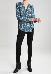 Teal långärmad blus med V-ringning, med ett mönster av vita hundar, ihop med svarta tajta jeans och svarta klackiga ankelboots.