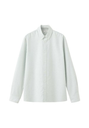 Jeune homme debout portant une chemise blanche à rayures boutonnée sur un t-shirt blanc, un pantalon marron à jambes larges, et des chaussures marron décontractées sur un fond uni.