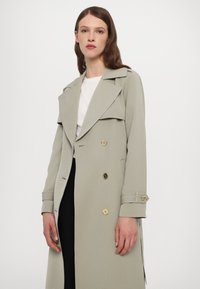 Double-breasted light grey coat with large lapels, gold buttons, and cuff detailing. Smooth fabric with a structured fit, worn over a white top.