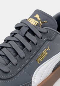 Grå sportskor med texturerat mocka och slätt läder på ovandelen, med ett vitt streck, grå snörning och guldiga logodetaljer.