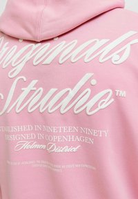Roze dukserica s kapuljačom koja sadrži bijeli natpis. Dizajn uključuje "Originals Studio" i podatke o podrijetlu i brendu. Glatka tekstura tkanine.