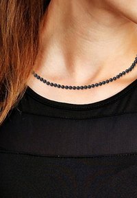 Collier de perles noires avec une texture lisse et uniforme, composé de perles sphériques espacées régulièrement. Porté au-dessus d'un haut noir sur un cou dénudé.