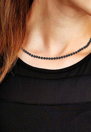 Collier de perles noires avec une texture lisse et uniforme, composé de perles sphériques espacées régulièrement. Porté au-dessus d'un haut noir sur un cou dénudé.