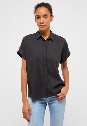 Frau trägt ein kurzärmeliges schwarzes Hemd mit Knöpfen und hellblaue Jeans, steht mit einer Hand in der Tasche vor einem einfarbigen Hintergrund.