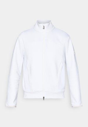 Veste blanche zippée avec un col haut, en tissu texturé, coutures plates et un petit logo sur la manche gauche. Présente des poches latérales.