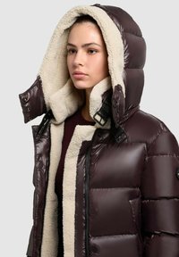 Giacca imbottita marrone scuro con una morbida fodera in pile color crema. Presenta un cappuccio, chiusura con zip e un materiale esterno lucido.