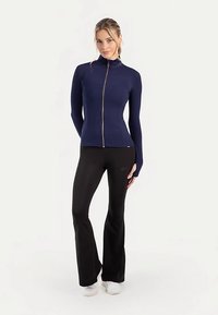 Veste zippée bleu marine avec col montant, manches longues et coupe ajustée, associée à un pantalon noir évasé et des chaussures blanches sur un fond neutre.