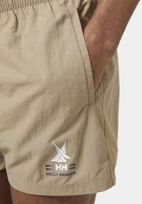 Bañador beige hecho de tejido ligero, con una cinturilla elástica, bolsillos laterales y un logo blanco con detalles azules en el dobladillo.