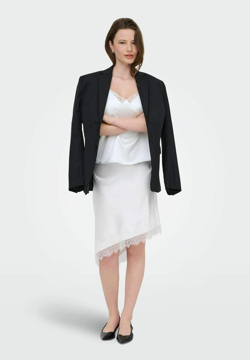 Femme portant une robe nuisette en satin blanc avec un ourlet en dentelle et un blazer noir drapé sur les épaules, debout avec les bras croisés et des talons noirs.