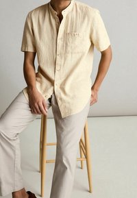 Chemise en lin beige clair à manches courtes avec une coupe décontractée, boutonnière sur le devant et une poche poitrine, associée à un pantalon de couleur claire.