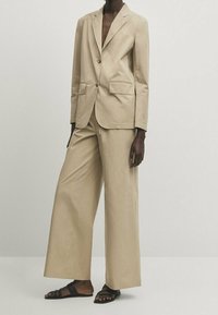 Beżowa, dopasowana marynarka z dwoma przednimi kieszeniami, zapinana na jeden guzik, zestawiona z luźnymi beżowymi spodniami i czarnymi sandałami na paski.