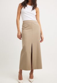 Beige, figurbetonter Maxikleid aus elastischem Stoff, mit einem Schlitz vorne für Bewegungsfreiheit. Kombiniert mit einem weißen Tanktop und High Heels.