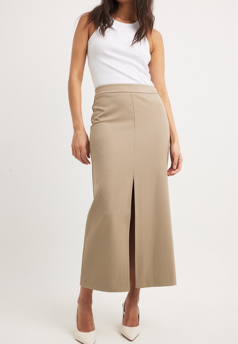 Beige, figurbetonter Maxikleid aus elastischem Stoff, mit einem Schlitz vorne für Bewegungsfreiheit. Kombiniert mit einem weißen Tanktop und High Heels.